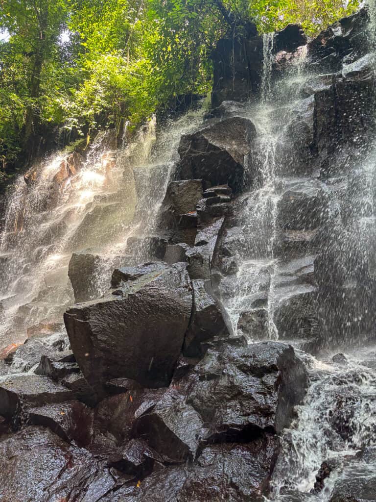 Kanto Lampo Waterfall cascading down dark volcanic rocks in a lush jungle setting, with sunlight filtering through the trees. A serene and photogenic stop on the Ubud waterfalls itinerary.