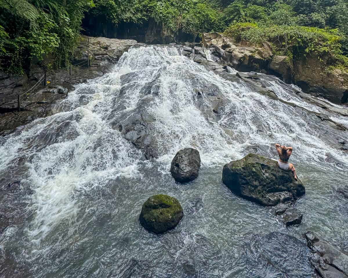 Goa Rang Reng Waterfall flowing in wide, gentle cascades over smooth dark rocks into a shallow river, surrounded by dense tropical greenery. A person sits on a large boulder at the base, adding scale to this tranquil Ubud waterfalls spot.