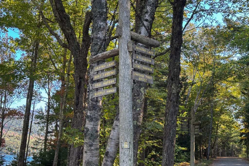 Wooden carriage road signpost in Acadia National Park showing directions to Around Lake, Seal Harbor, Bubble Pond, and Jordan Pond House. The sign is mounted to a tree in a forested trail intersection, with sunlight filtering through green and autumn-tinged leaves.