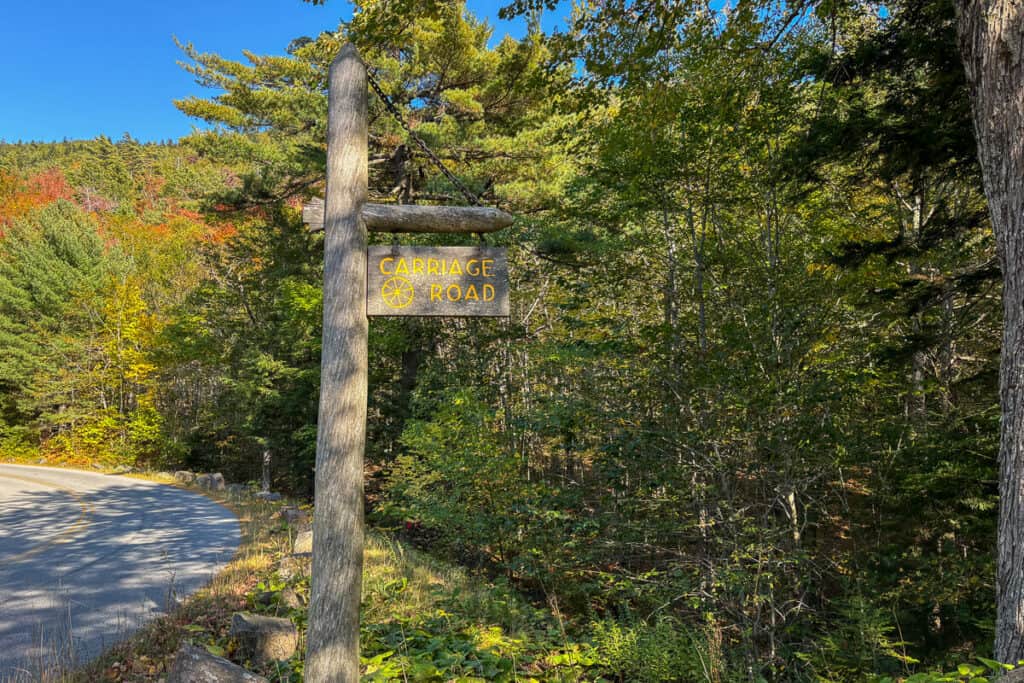Wooden Carriage Road sign hanging from a post near a forested trailhead in Acadia National Park. The sign features yellow text and a wagon wheel symbol, with a curving paved road and autumn foliage in the background.