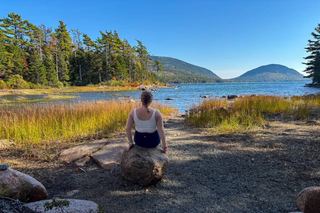 Woman with a braid sits on a rock facing Eagle Lake in Acadia National Park, surrounded by tall grasses and framed by forested hills and a clear blue sky. The peaceful lakeside view highlights the natural beauty along the carriage roads.