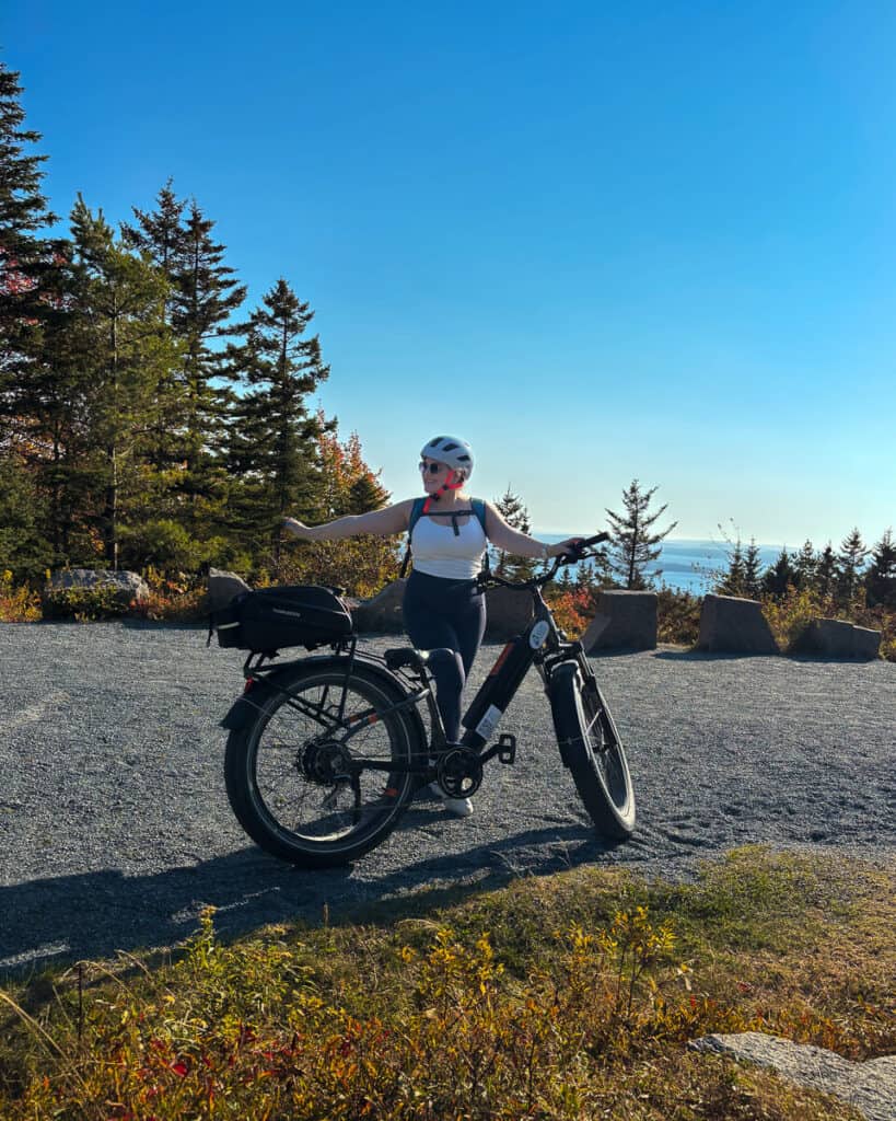 Woman posing with an e-bike at a scenic overlook on Acadia’s carriage roads, pointing toward the ocean in the distance. She’s wearing a helmet and backpack, surrounded by evergreens, autumn shrubs, and a clear blue sky.