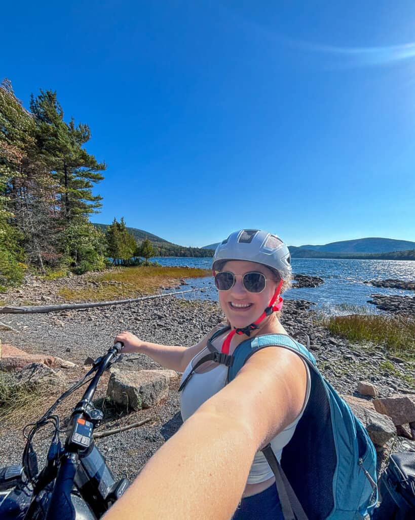 Woman smiling in a selfie while holding an e-bike near the rocky shore of Eagle Lake in Acadia National Park. She's wearing a helmet, sunglasses, and a backpack, with clear blue skies and forested hills in the background along the carriage roads