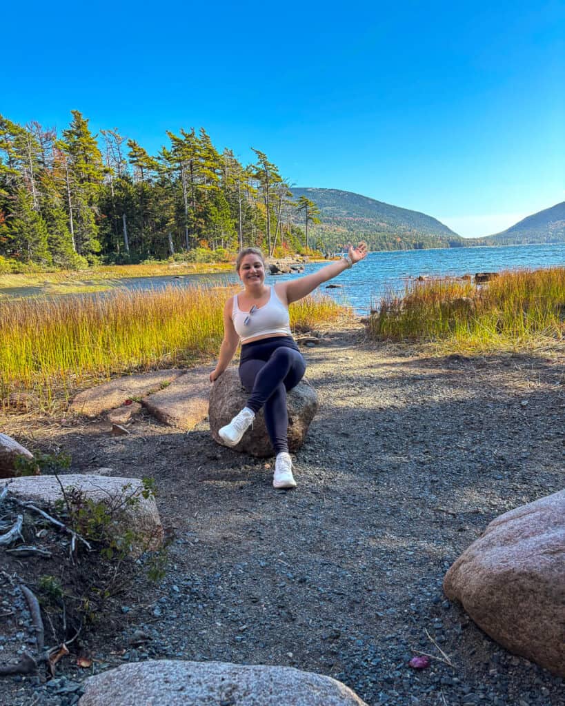Woman sitting on a large rock at the edge of Eagle Lake in Acadia National Park, smiling and gesturing toward the water and surrounding mountains. She's dressed in activewear, with golden grasses, pine trees, and a bright blue sky adding to the scenic backdrop along the carriage roads.
