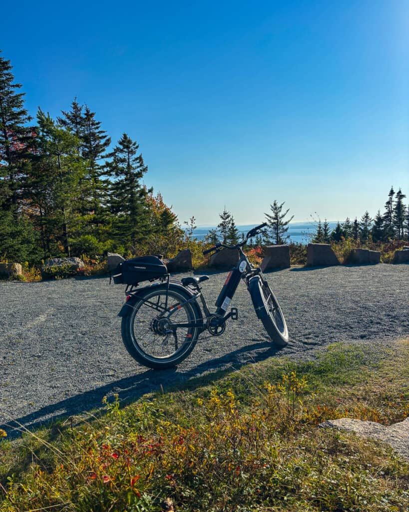 Electric bike parked on a gravel pull-off along Acadia National Park’s carriage road, overlooking a scenic view of forested hills and the Atlantic Ocean under a clear blue sky. Captures a peaceful stop during an Acadia e-bike ride through fall foliage.