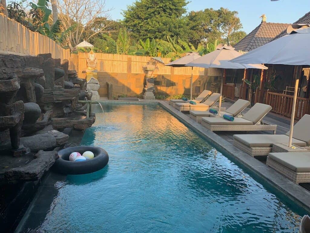 Pool area at Royal Cottages Nusa Lembongan featuring cushioned sun loungers with umbrellas, a stone fountain wall, and tropical landscaping. A black float with beach balls rests in the water, capturing the relaxed vibe of this top accommodation option when deciding where to stay in Nusa Lembongan.