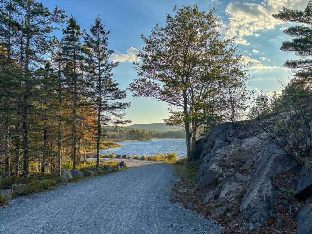 Gravel carriage road in Acadia National Park curves gently toward a lake, with trees lining both sides and late afternoon sunlight casting a golden glow on the water and trail. A rocky ledge rises on the right as the view opens to a peaceful lakeside scene under a partly cloudy sky.