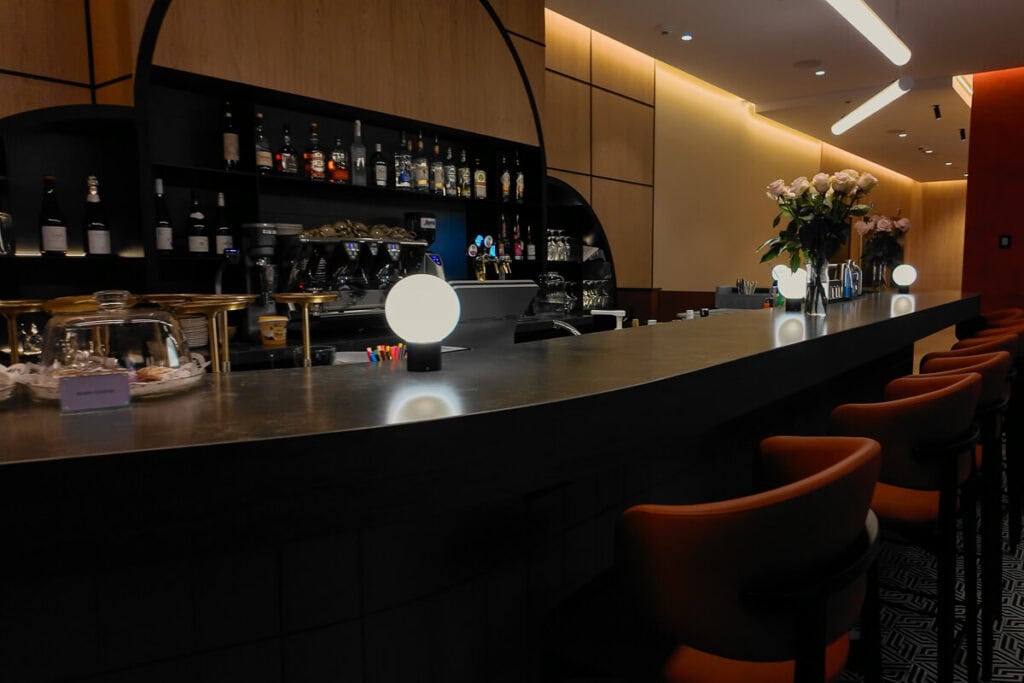 Modern bar area at Hyatt Place Tallinn featuring a sleek countertop, glowing orb lights, leather barstools, a flower arrangement, and a well-stocked back bar with spirits and coffee equipment.