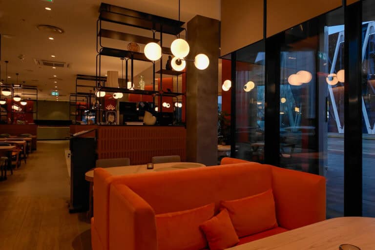 Chic dining area at Hyatt Place Tallinn with modern globe lighting, bold orange booth seating, wood tables, and large windows offering a street view of the city’s contemporary architecture.