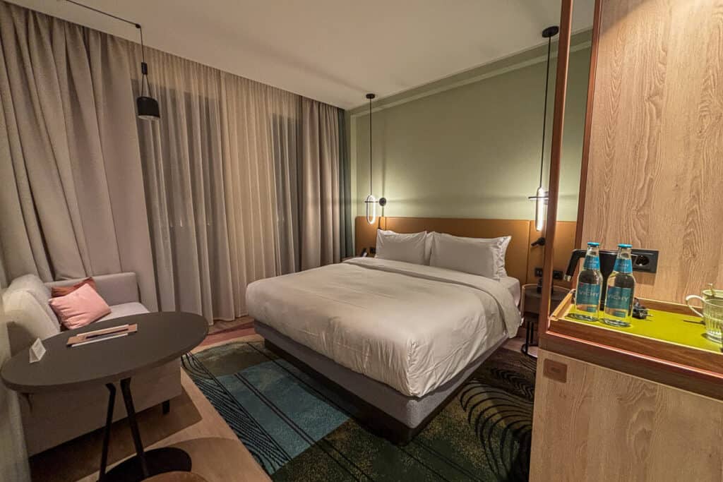 Stylish room at Hyatt Place Tallinn with a king-sized bed, hanging pendant lights, a lounge area with a sofa and table, and complimentary Vichy water bottles on a wooden refreshment counter.
