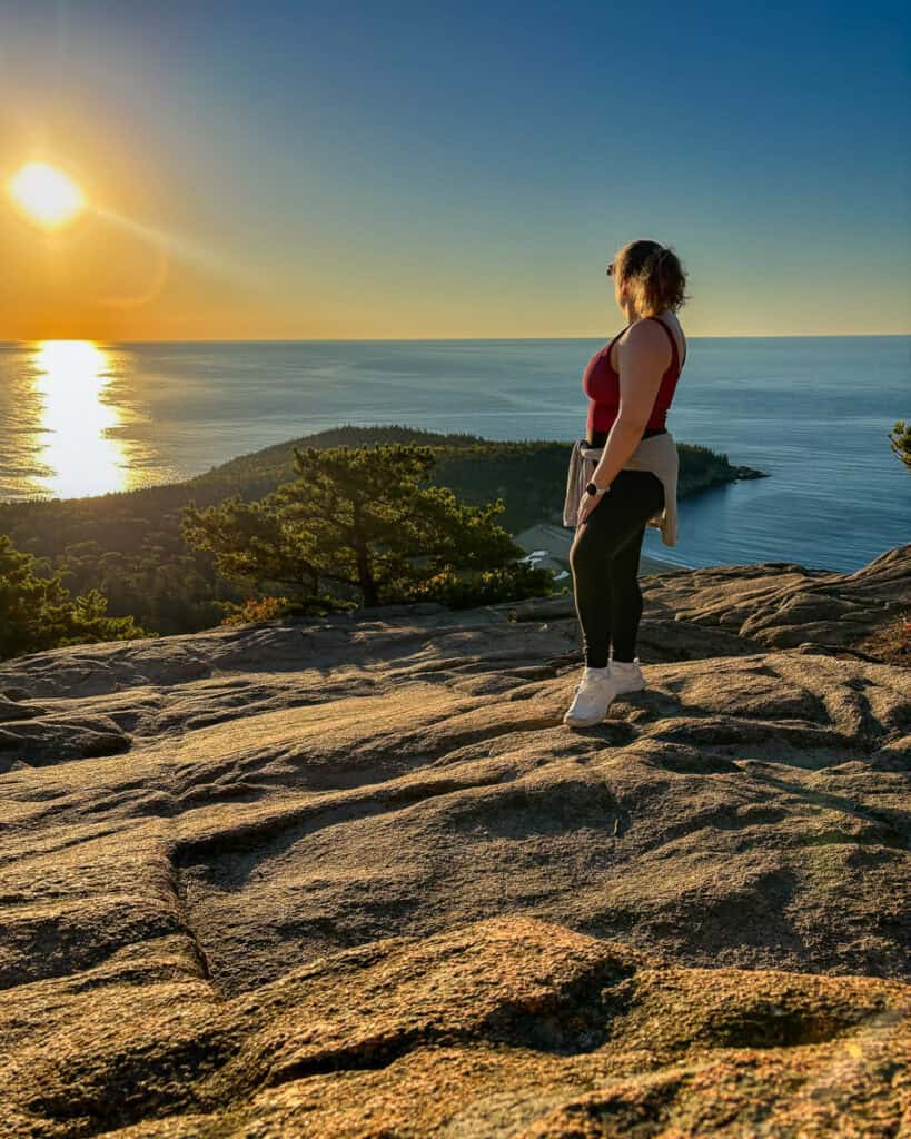 A woman stands on sunlit granite after completing the Beehive Trail in Acadia National Park, gazing at the sunrise glowing over the Atlantic Ocean. The moment captures both achievement and stillness, symbolizing the deeper life lessons from travel found on a New England fall road trip.