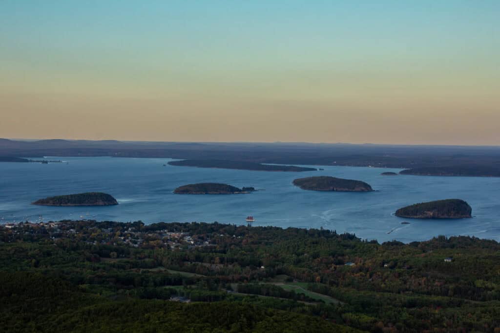 A wide view from Cadillac Mountain captures the early hues of sunset over the islands and coastal town below, with a tall ship sailing through the bay.