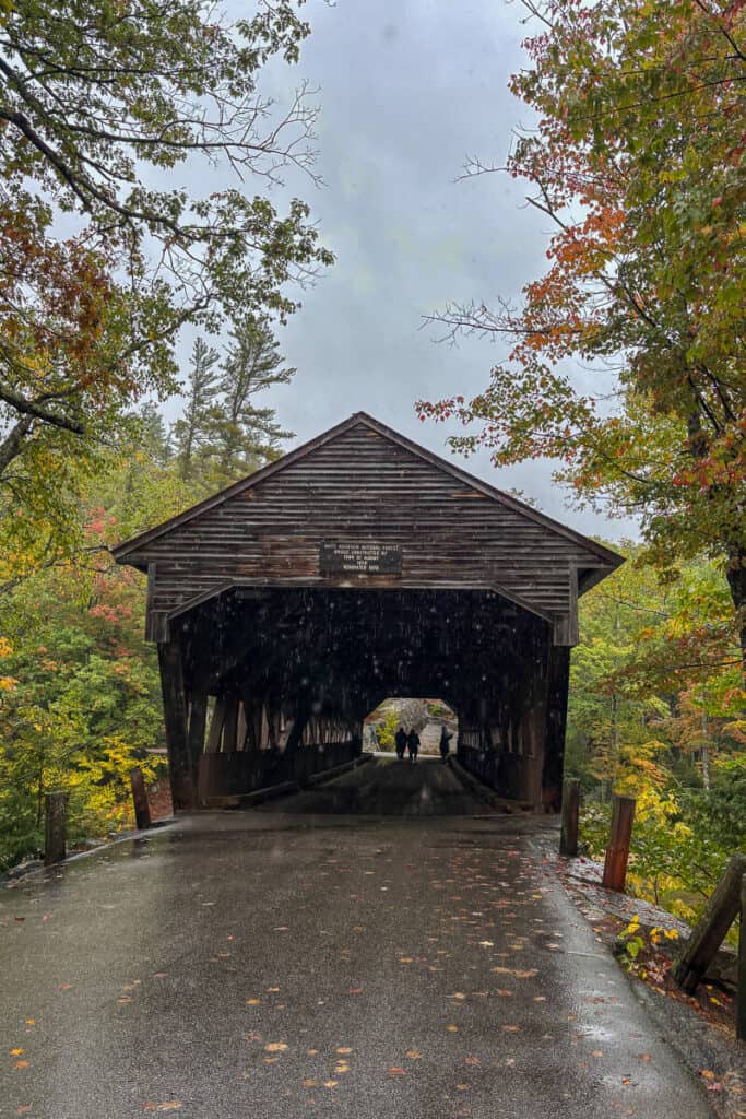 A few people walk through a wooden covered bridge along the Kancamagus Highway in New Hampshire, as rain begins to fall and autumn leaves cling to the wet pavement.