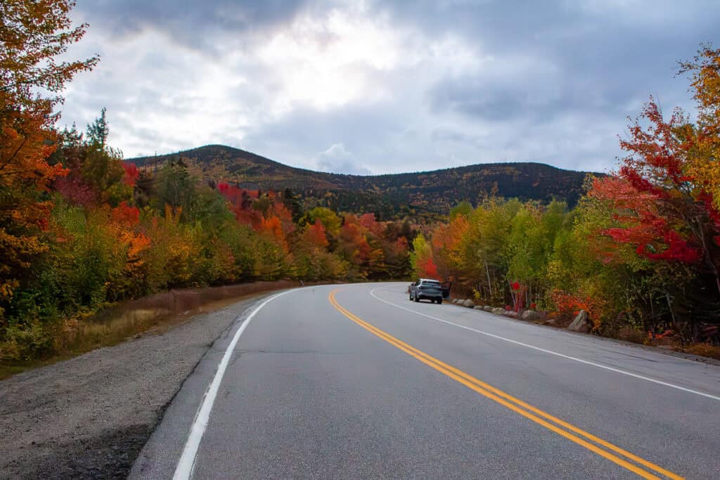 A car stopped along a winding stretch of the Kancamagus Highway in New Hampshire, framed by vibrant fall foliage under a moody sky.
