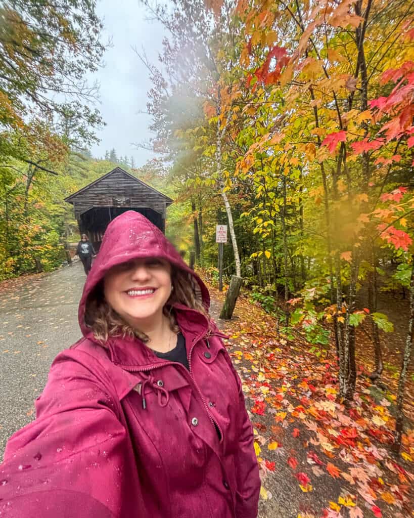 A smiling woman in a rain-soaked maroon raincoat takes a selfie near a covered bridge in New Hampshire's White Mountains, surrounded by vibrant fall foliage and wet leaves.
