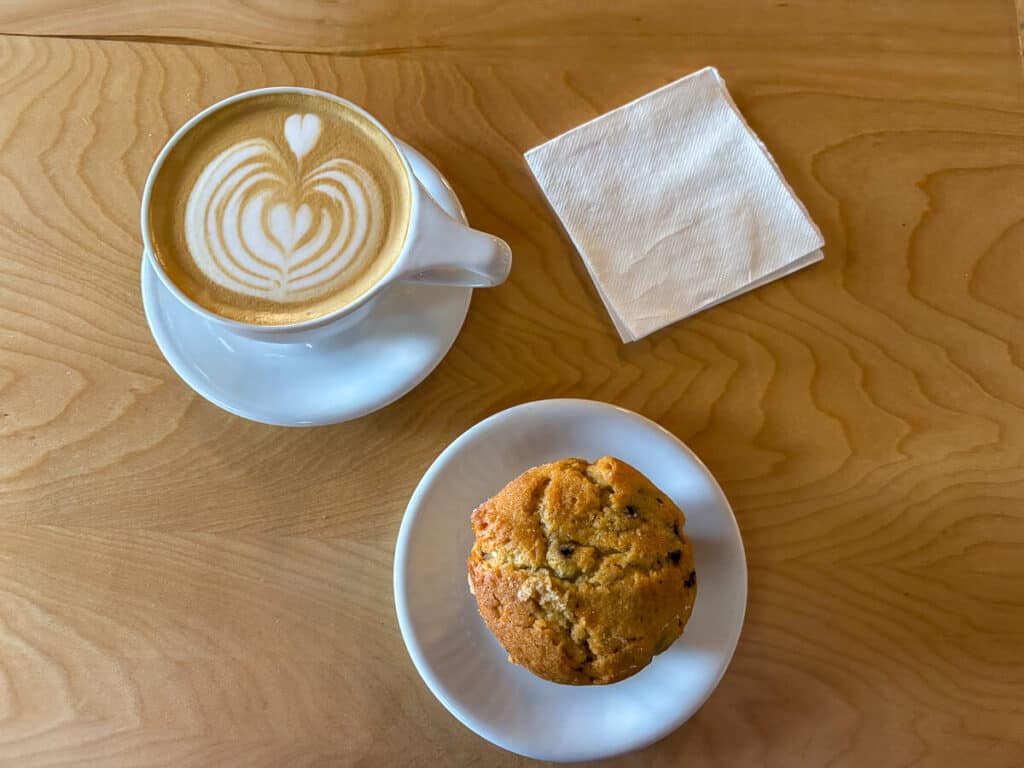 A warm latte with heart-shaped foam art and a blueberry muffin sit on a wooden table beside a stack of napkins in a New Hampshire coffee shop. The inviting scene offers a quiet pause and simple comfort, echoing life lessons from travel like slowing down and savoring the moment.