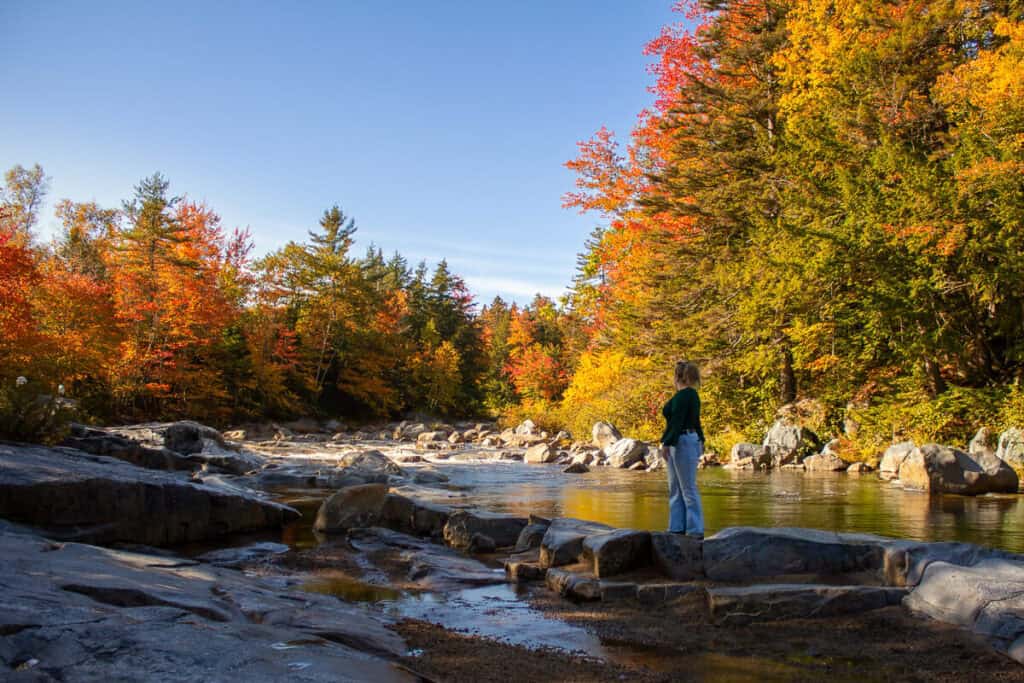 A woman stands on river rocks along the Kancamagus Highway in New Hampshire, surrounded by brilliant fall foliage reflecting in the calm water.