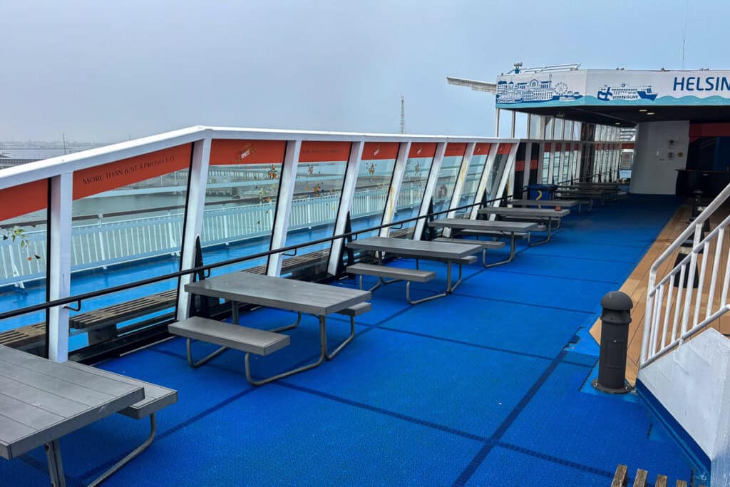 Outdoor seating area on the Eckerö Line ferry during a Tallinn to Helsinki crossing, with picnic-style tables, bright blue flooring, and glass panels displaying the slogan “More than just a prosecco.” The deck is empty under an overcast sky.