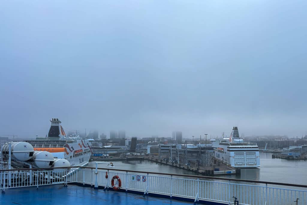 Foggy morning view of Tallinn harbor from the deck of a ferry, with two Tallink ships docked and the city skyline barely visible in the mist. A typical departure scene for the Tallinn to Helsinki ferry route.