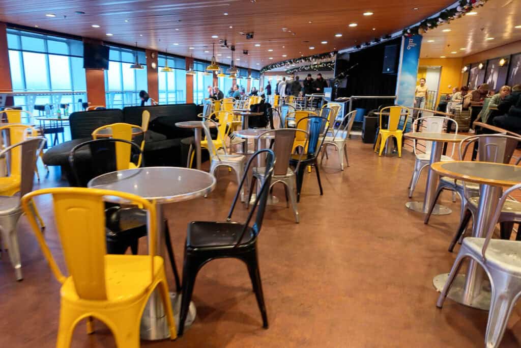 Bright and casual seating area inside a Tallinn to Helsinki ferry, featuring metal tables and a mix of yellow, black, and silver chairs. Large windows line the side, offering sea views during the crossing.