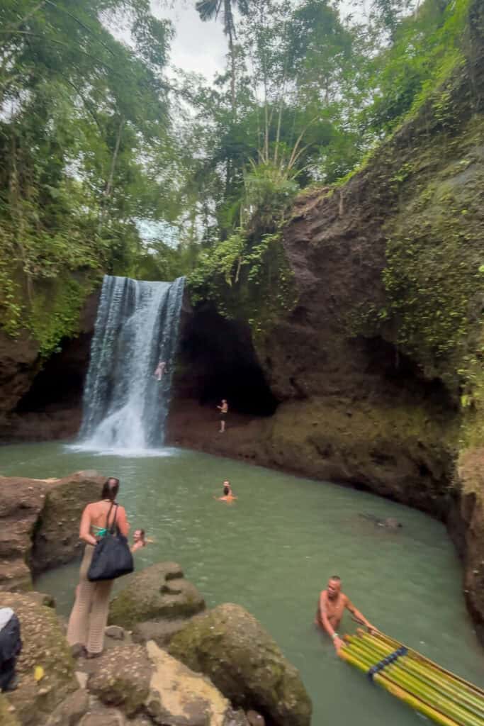 Visitors swimming and relaxing at the base of a secluded jungle waterfall with a steady stream flowing into a turquoise pool, surrounded by mossy cliffs. This scenic spot is part of the Ubud waterfalls day trip in Bali.