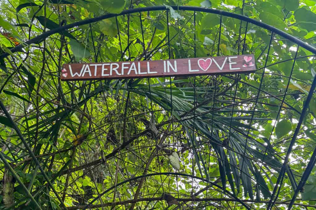 A wooden sign reading “Waterfall in Love” with hand-painted hearts, hanging from a metal archway surrounded by dense tropical foliage. A whimsical photo spot along the Ubud waterfalls trail.