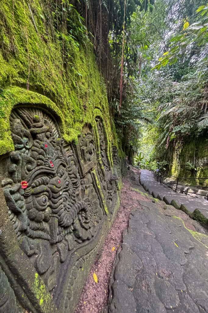 Moss-covered stone carvings of Balinese mythological figures line the stairway leading through the jungle to Goa Rang Reng Waterfall. Intricate details and scattered red flower offerings add spiritual ambiance to this Ubud waterfalls destination