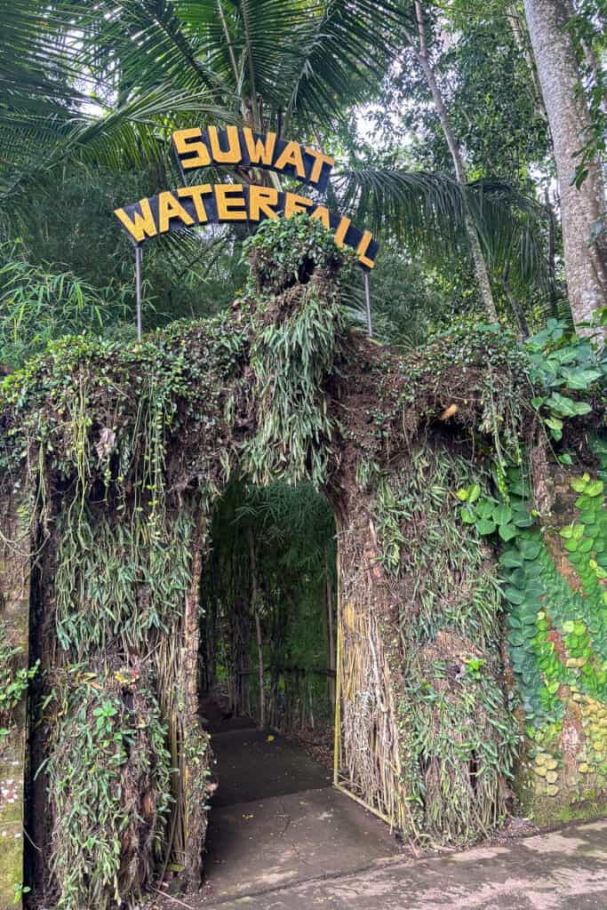 Entrance to Suwat Waterfall framed by a rustic archway made of vines, roots, and tropical plants, with a bold yellow sign above. A lush and inviting start to one of the hidden gems on the Ubud waterfalls trail.