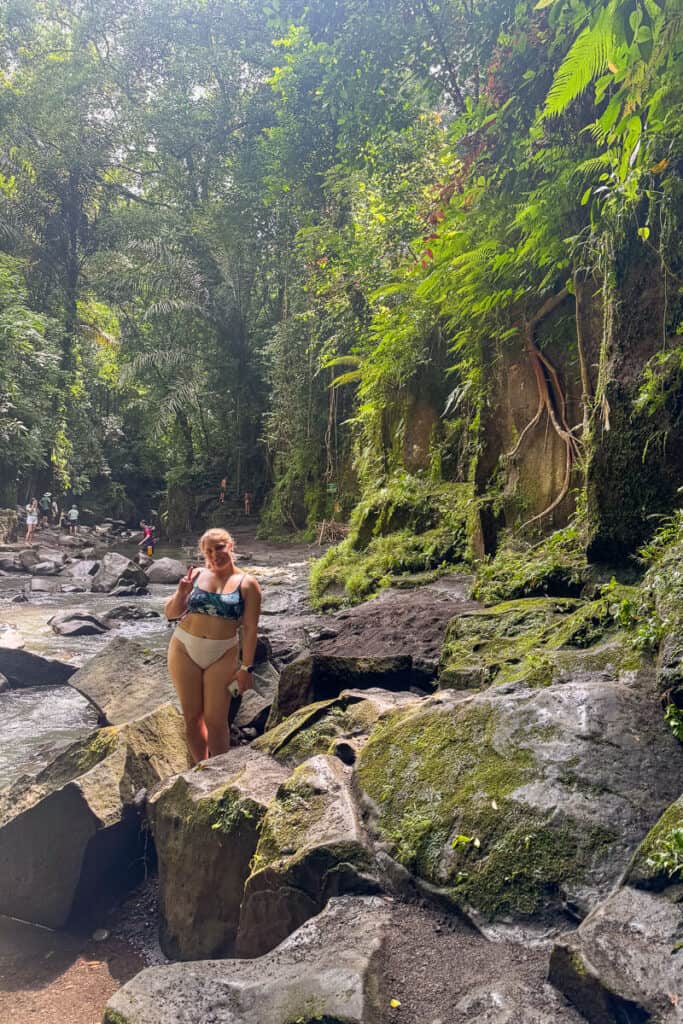 Traveler posing with a peace sign among moss-covered rocks and dense jungle foliage at the base of Kanto Lampo Waterfall in Bali. A peaceful, shaded stop along the Ubud waterfalls route.