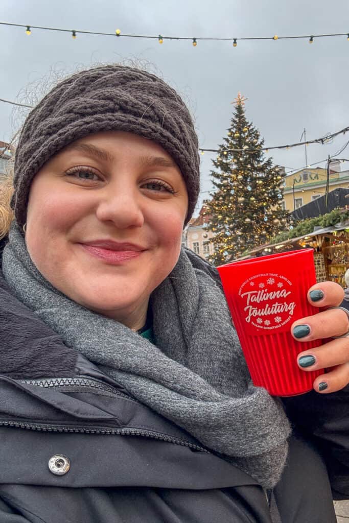 Woman holding a red cup reading "Tallinna Jõuluturg" at the Tallinn Christmas market with a decorated tree glowing behind her, enjoying a warm drink.