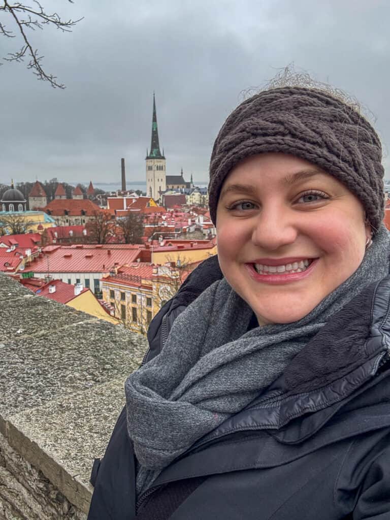 Woman in a knit headband and scarf smiling at a Tallinn Old Town viewpoint with red rooftops and St Olaf’s Church behind her, capturing a scenic moment during one day in Tallinn.