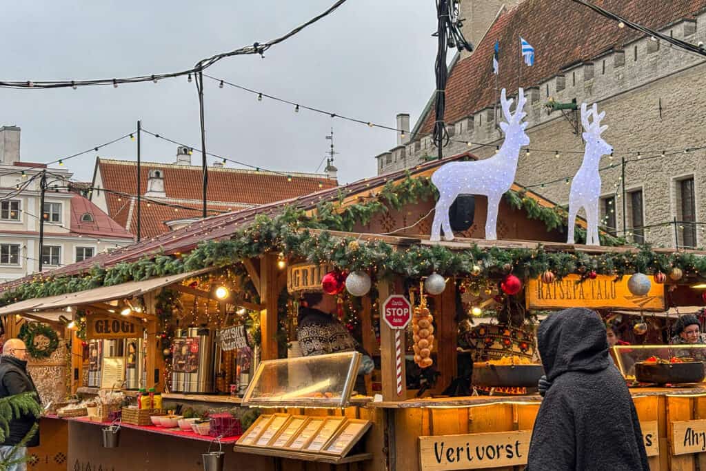 Festive Christmas market stall in Tallinn decorated with garlands and lights with glowing reindeer figures on the roof and people ordering hot food, a cozy winter experience during one day in Tallinn.