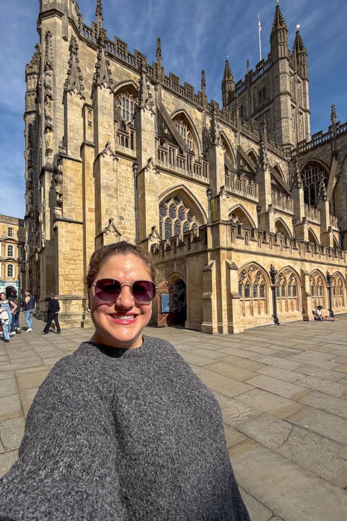 Woman smiling in sunglasses in front of Bath Abbey’s detailed stone facade and gothic windows on a sunny day, a must see landmark on a 2 day Bristol and Bath itinerary from London.