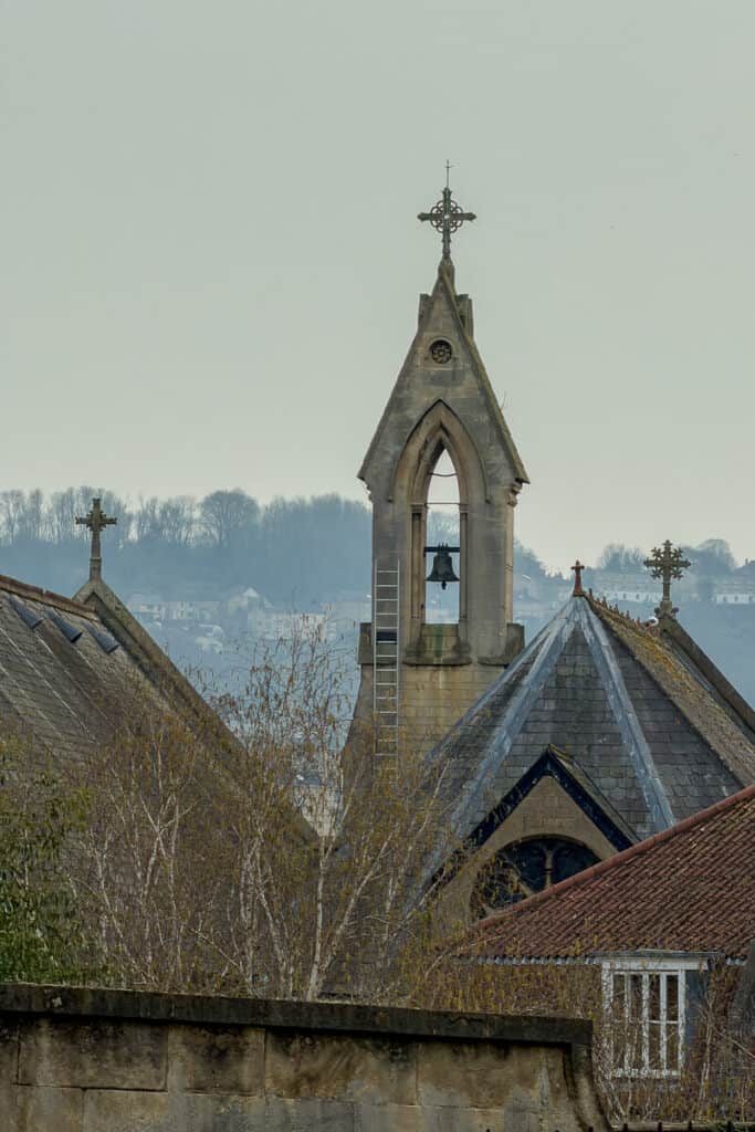 Stone church bell tower with a cross at the top rising above rooftops in Bath on a misty day, with trees and hillside homes in the background, a quieter view on a 2 day Bristol and Bath itinerary from London.