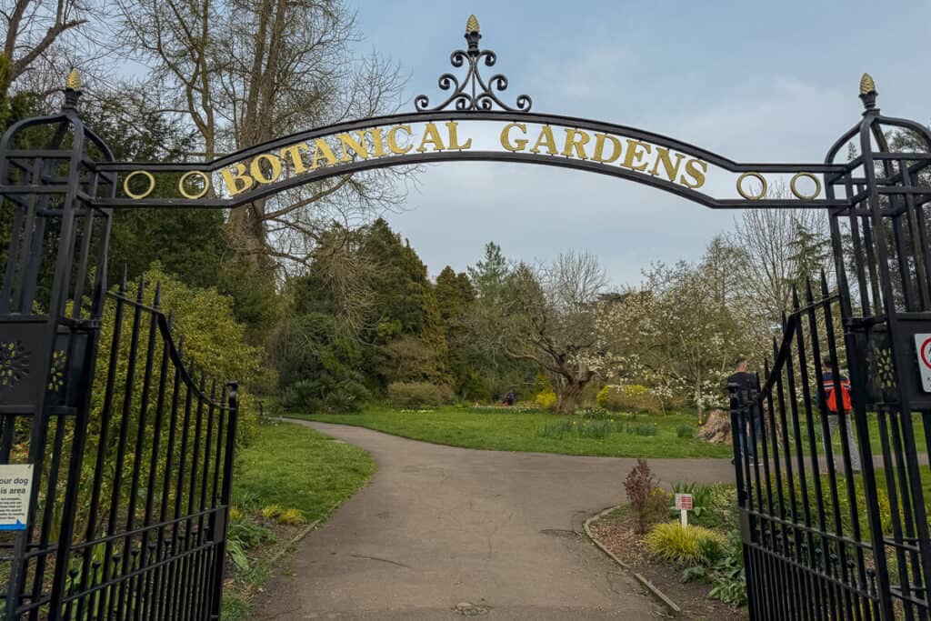 Entrance gate with gold lettering reading “BOTANICAL GARDENS” opening onto a winding path surrounded by greenery and blooming trees in Bath, a peaceful stop on a 2 day Bristol and Bath itinerary from London.