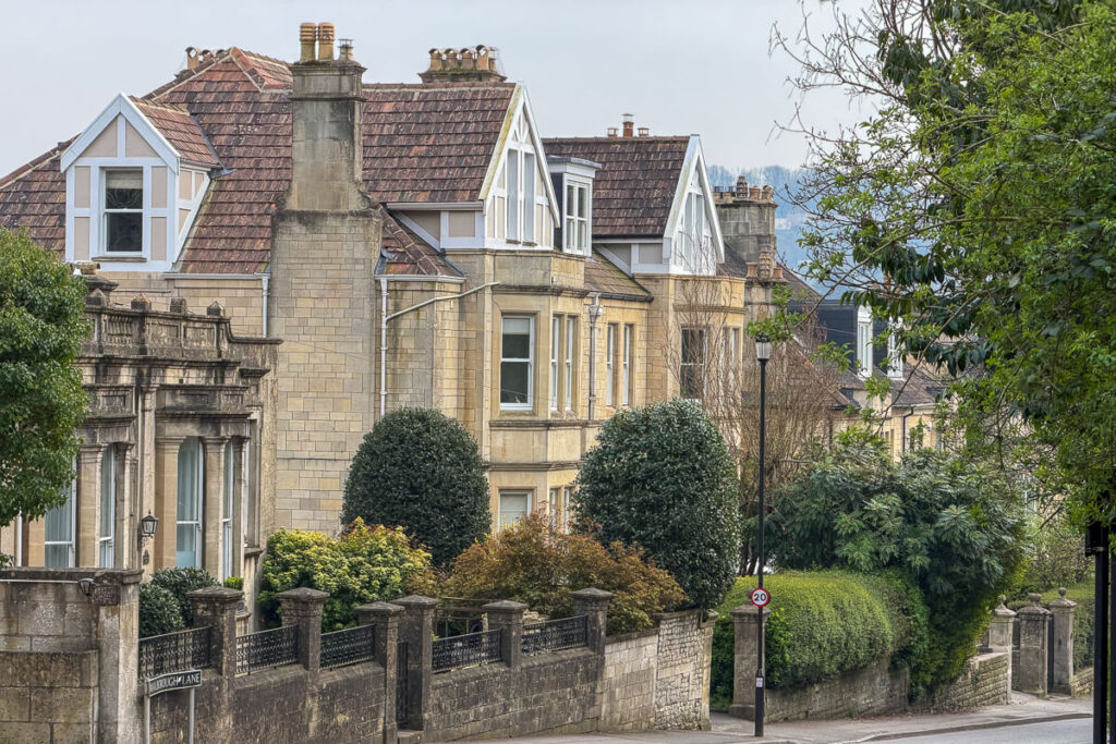 Row of elegant stone townhouses with bay windows and tiled roofs along a quiet residential street in Bath, surrounded by greenery, showing the charming architecture on a 2 day Bristol and Bath itinerary from London.