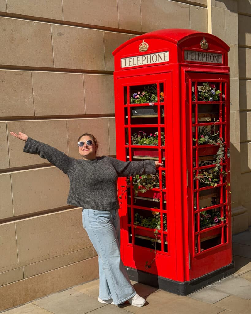Woman posing with arms outstretched beside a bright red British telephone booth filled with flowers against a stone wall in Bath, a fun photo spot on a 2 day Bristol and Bath itinerary from London.