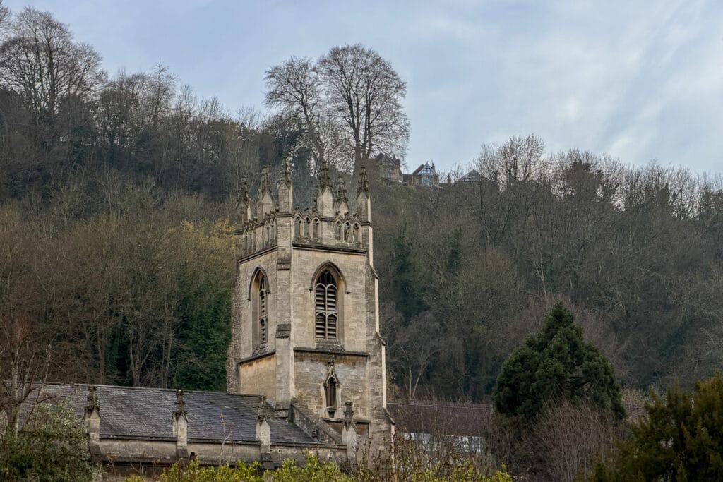 Historic stone church tower with gothic details set against a wooded hillside in Bath under an overcast sky, showing a quieter scenic side of a 2 day Bristol and Bath itinerary from London.