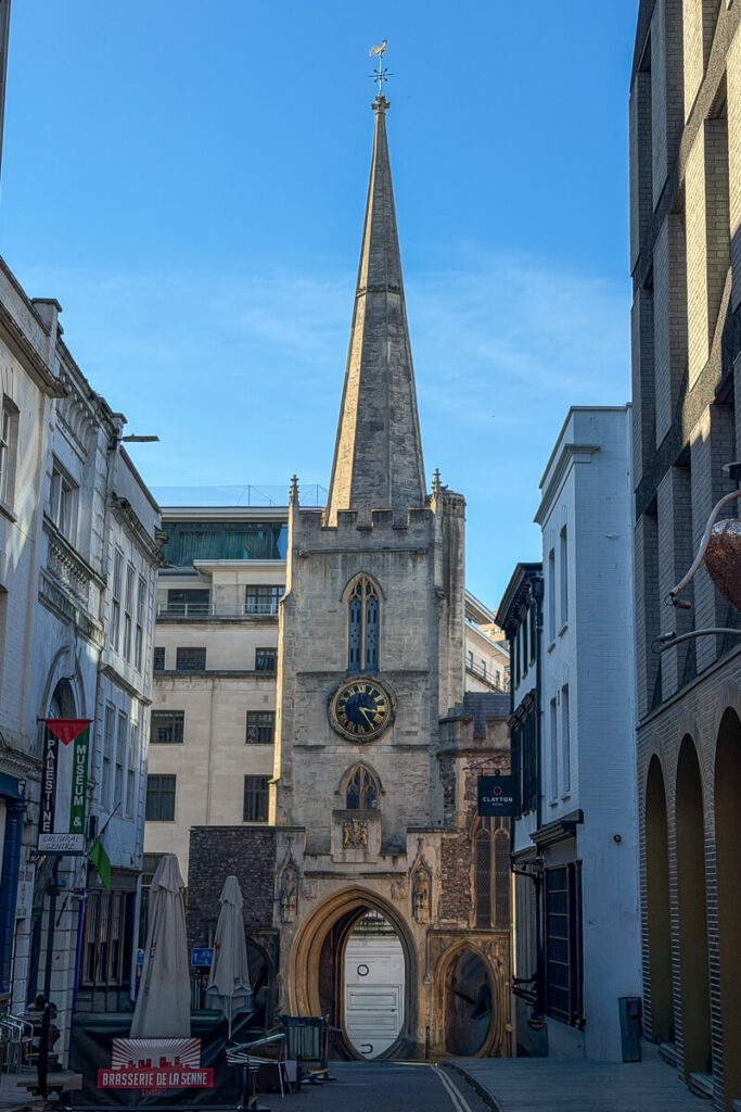 Tall church spire with a clock tower rising above a narrow street in Bristol surrounded by historic and modern buildings under a clear blue sky, a striking landmark on a 2 day Bristol and Bath itinerary from London.