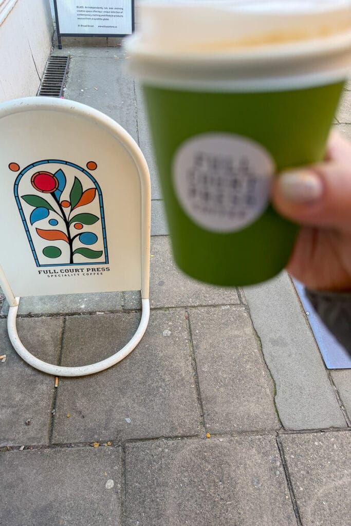 Hand holding a takeaway coffee cup in front of a street sign reading “FULL COURT PRESS SPECIALITY COFFEE” with a colorful floral logo in Bristol, highlighting a local cafe stop on a 2 day Bristol and Bath itinerary from London.