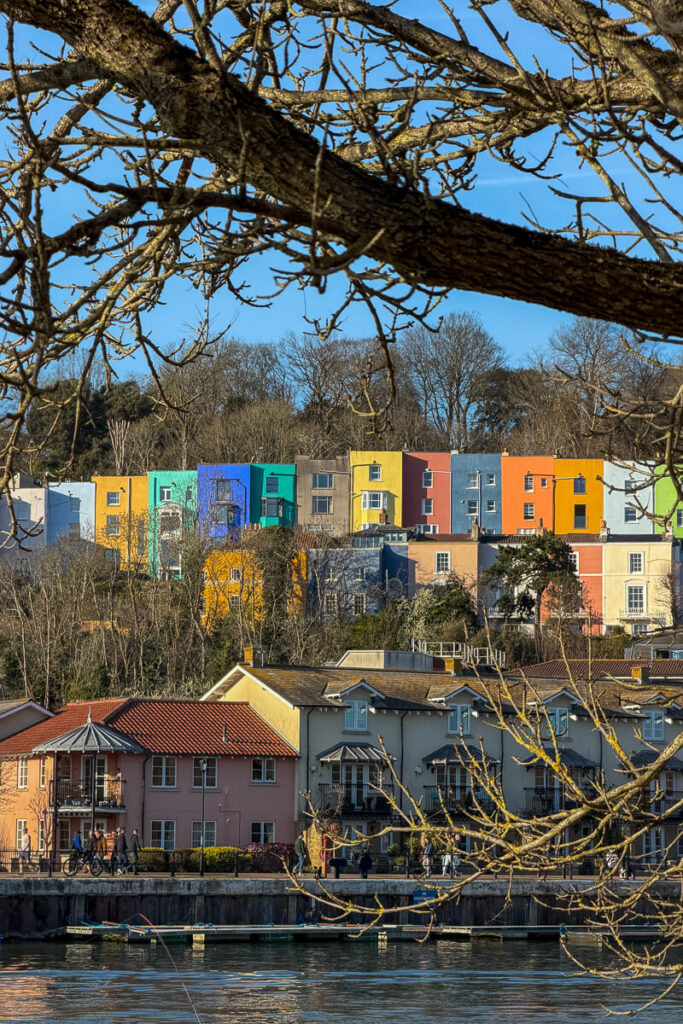 Colorful row of bright painted houses on a hillside in Bristol overlooking the waterfront with branches framing the view, a vibrant scene on a 2 day Bristol and Bath itinerary from London.