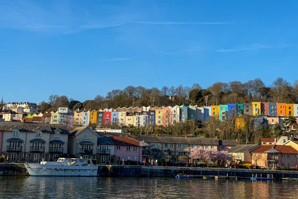 Colorful row of pastel houses lining the waterfront in Bristol with boats docked along the harbor under a clear blue sky, a vibrant stop on a 2 day Bristol and Bath itinerary from London.
