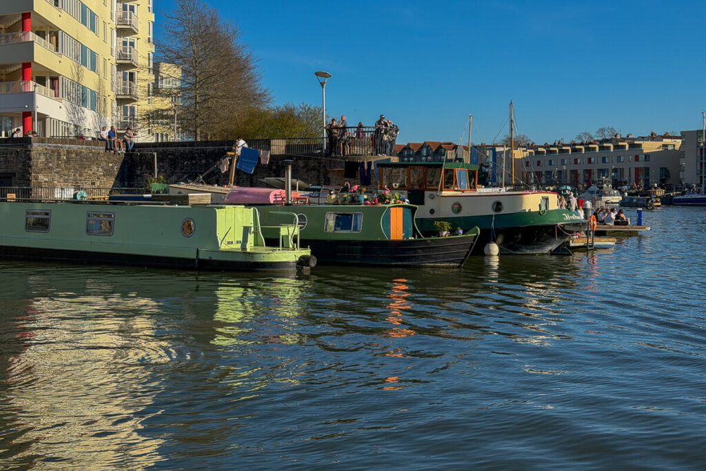 Houseboats moored along Bristol’s harbor with people relaxing on the waterfront and modern apartments in the background on a sunny day, showing the laid back side of a 2 day Bristol and Bath itinerary from London.