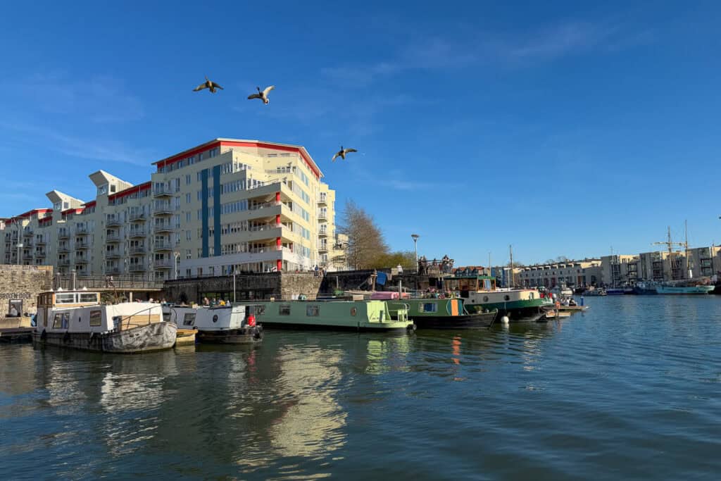Boats and houseboats along Bristol’s harbor with modern waterfront apartments and people gathered along the quay under a bright blue sky, with birds flying overhead, capturing the lively atmosphere of a 2 day Bristol and Bath itinerary from London.