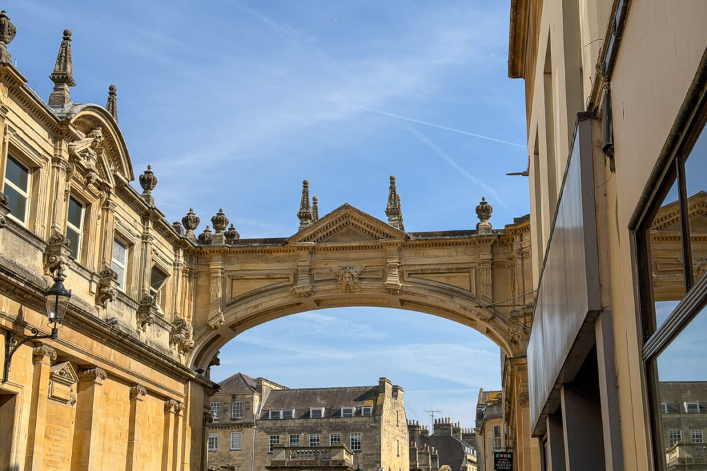 Ornate stone archway of Pulteney Bridge in Bath framed by historic buildings under a clear blue sky, a must see stop on a day trip to Bath from London.
