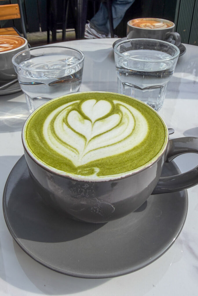 Matcha latte with heart shaped foam art on a cafe table alongside water glasses and coffee, a relaxing stop to include on a Bath day trip.