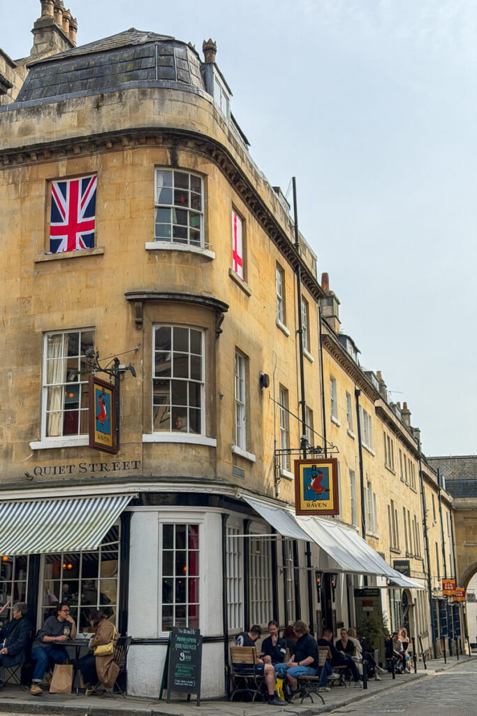 Historic honey colored buildings along Quiet Street in Bath with The Raven pub and people sitting outside, capturing the lively atmosphere of a Bath day trip.