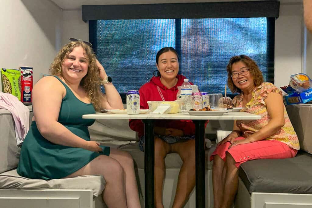 Three women sitting at a dinette table inside an RV, smiling with snacks and drinks spread out.