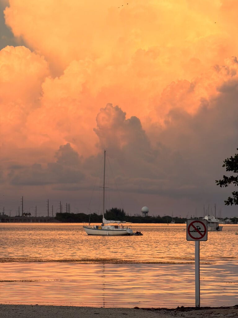 Dramatic orange storm clouds over a harbor at sunset, with a sailboat and a no-swimming sign in the foreground.